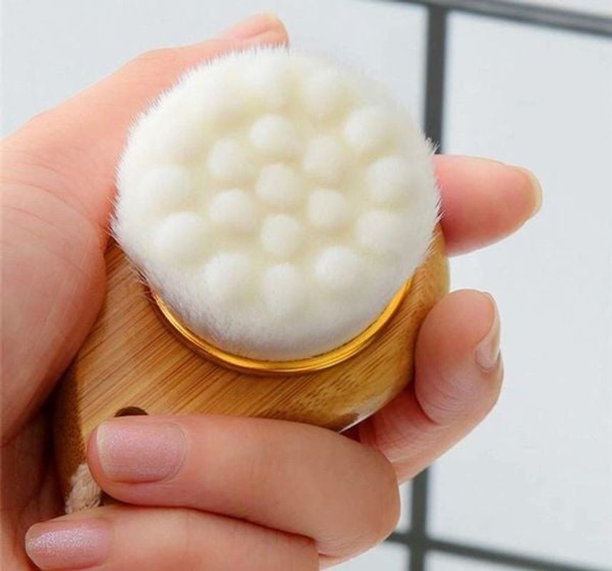 Hand holding a wooden facial cleansing brush with a white bristle head.